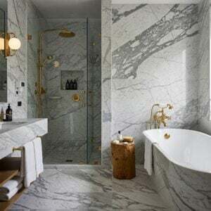 A luxurious boutique hotel bathroom featuring elegant white marble walls and flooring with dramatic gray veining. The space is adorned with gold fixtures, a freestanding bathtub, a glass-enclosed shower, and a rustic wooden stool, creating a refined yet inviting ambiance.